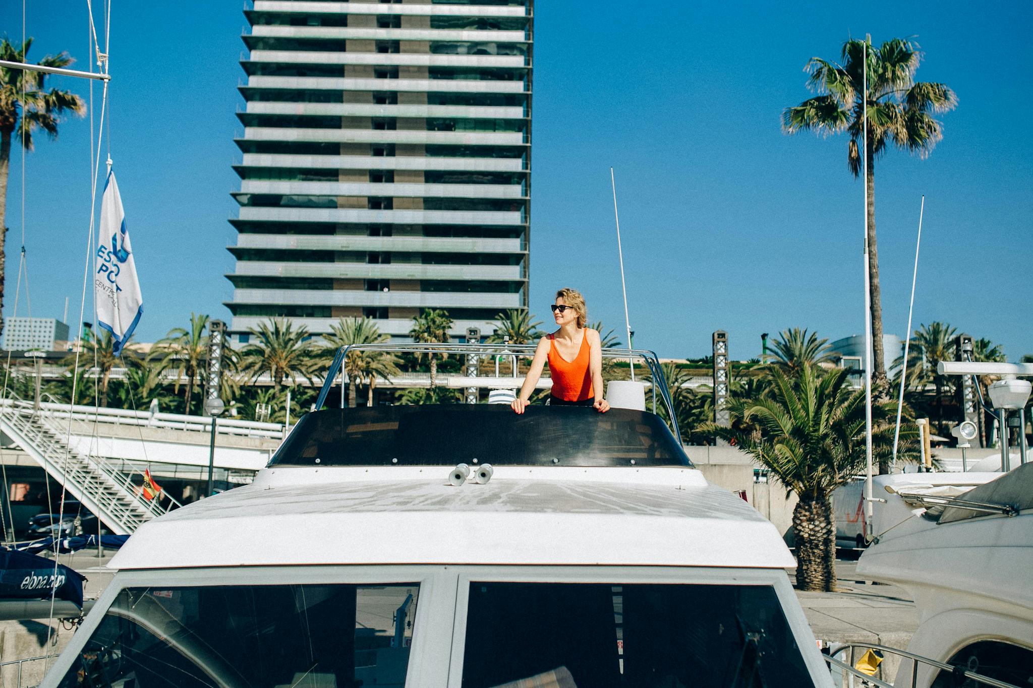 Woman enjoying a sunny day on a yacht docked by palm trees and modern buildings.